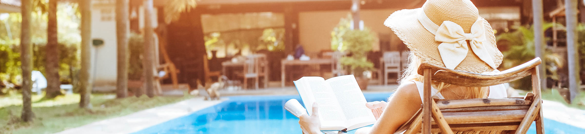 Frau liest Buch am Pool mit Ferienhaus im Hintergrund